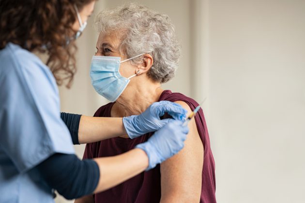 oudere vrouw met mondkapje op krijgt injectie van zorgmedewerker